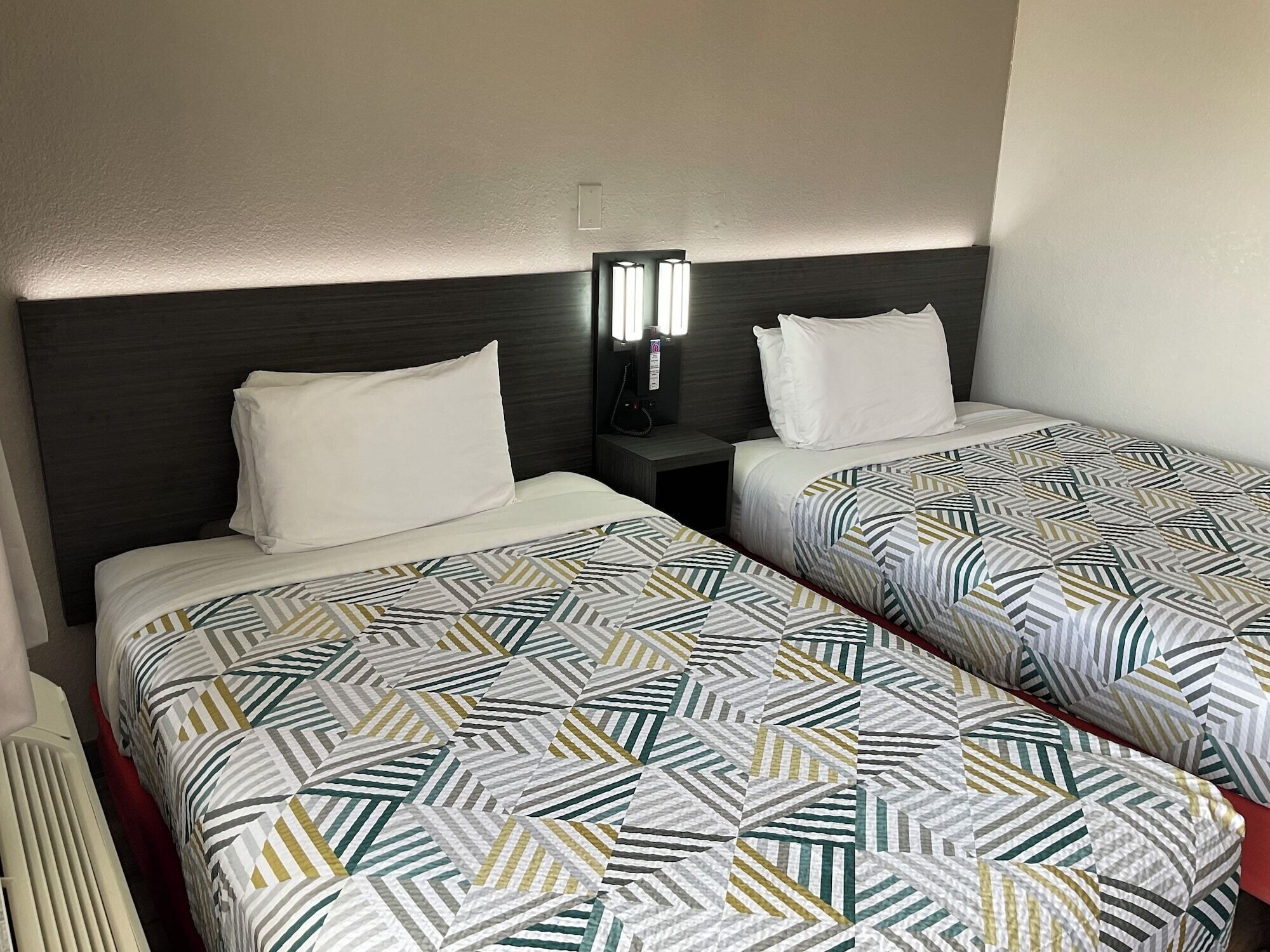 A hotel room with two beds, each with patterned bedding, a bedside table with a lamp, and a wall-mounted light fixture.