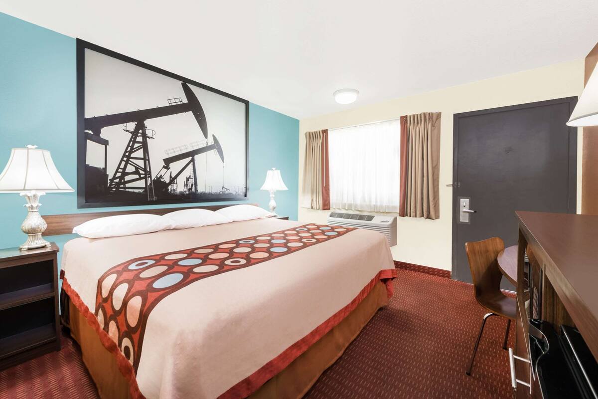 A hotel room with a large bed, two bedside tables with lamps, a desk with a chair, a window with curtains, and a door.