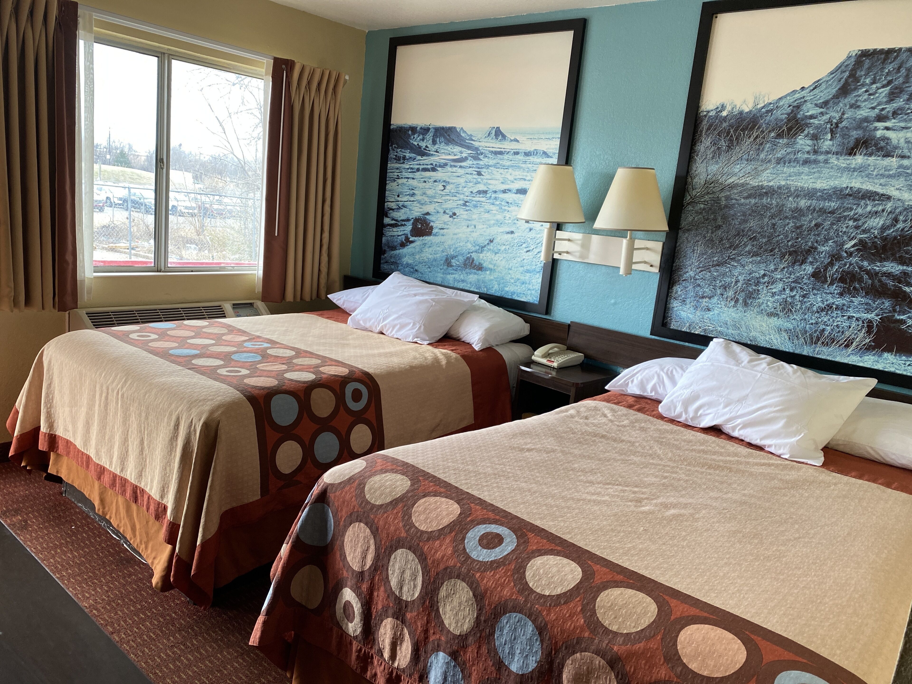 Quarto de hotel com duas camas, uma janela grande com cortinas, um quadro na parede e um abajur na mesa de cabeceira.