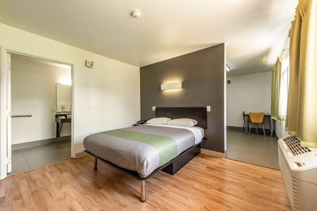 A single bed with a headboard, a bedside table, a mirror, and a window with curtains.
