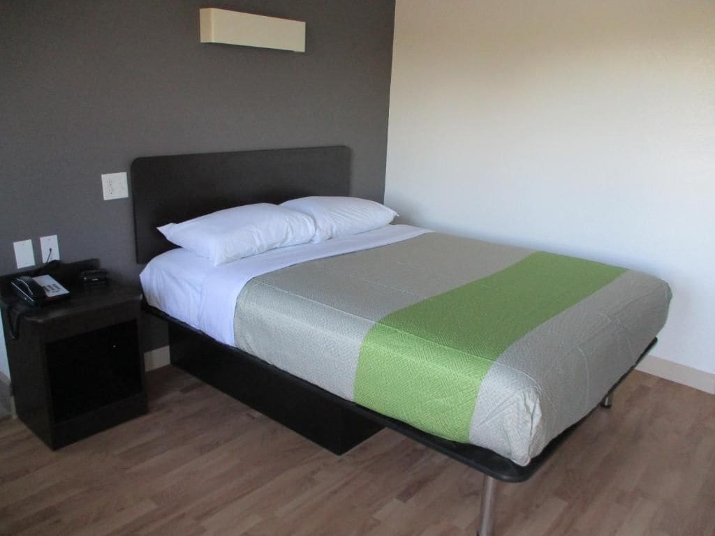 Un lit simple avec un matelas gris et blanc, une touche de vert, une tête de lit noire, une table de chevet avec un téléphone et un luminaire fixé au mur.