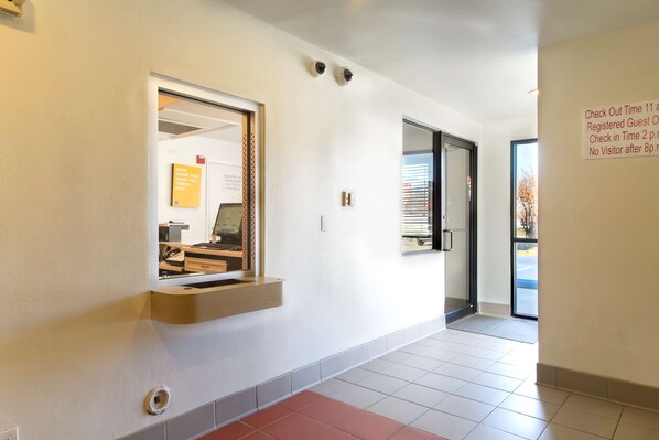 A clean, modern hallway with a reception desk, a check-in/out sign, and a security camera. - Motel 6 Capitol Heights, MD - Washington (Capitol Heights)