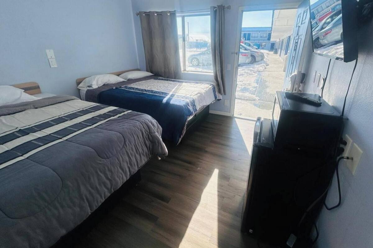 A hotel room with two beds, a window with curtains, a desk with a computer, and a view of the outside parking area.