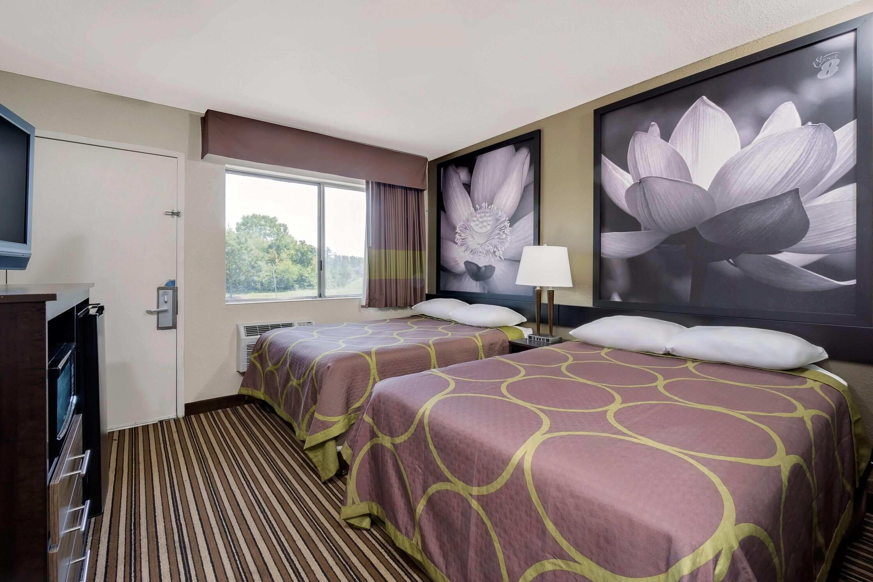 Una habitació d'hotel amb dos llits, una televisió, una finestra amb cortines i un quadre gran amb flors a la paret.