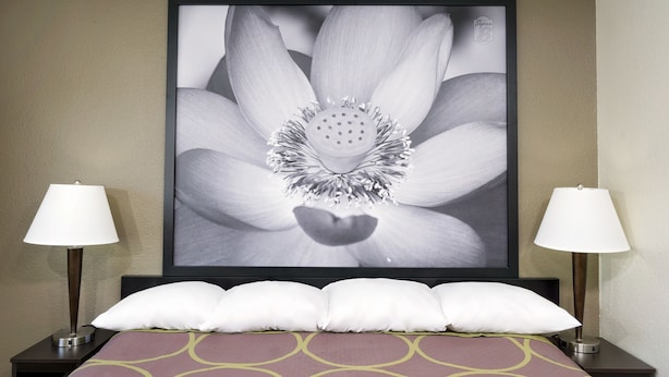 Una habitació d'hotel amb una gran obra d'art floral emmarcada a la paret, un llit amb un estriatge i dos llençols, i un conjunt de coixins blancs.