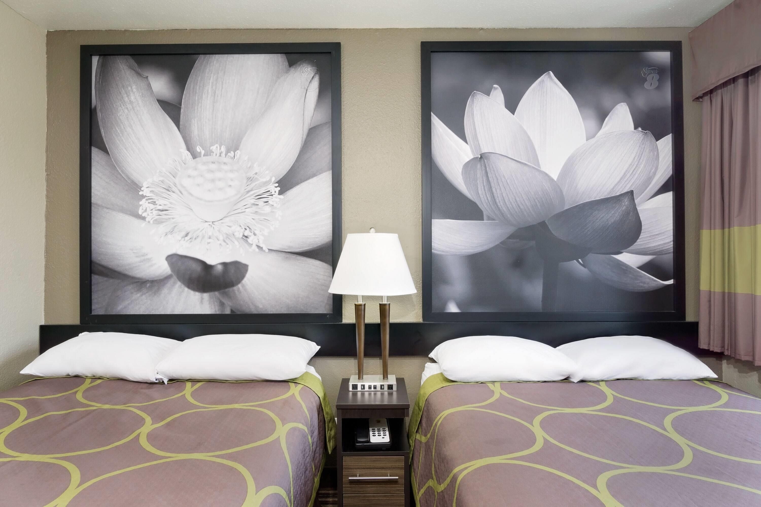 A hotel room with two beds, a nightstand with a lamp, and two large framed floral artworks on the wall.