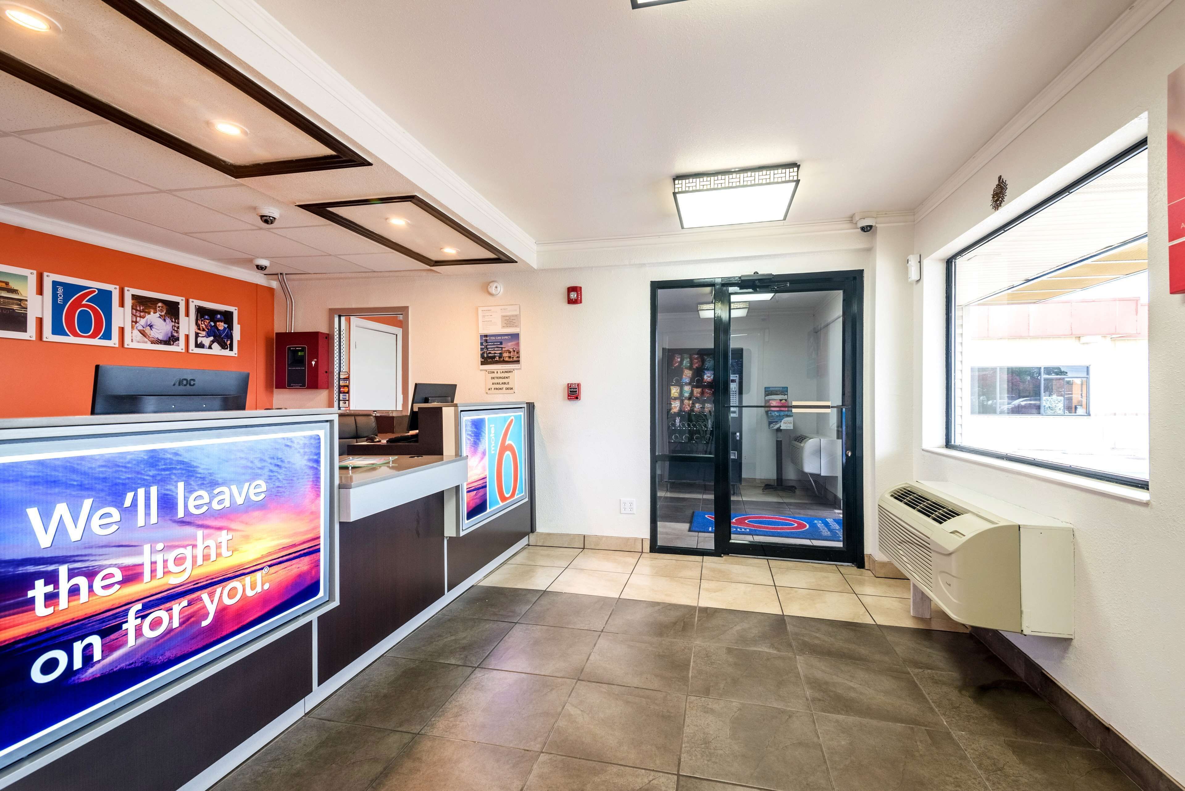 Zona de recepción de hotel con un letrero digital, mostrador de recepción y puerta de vidrio que da al vestíbulo.