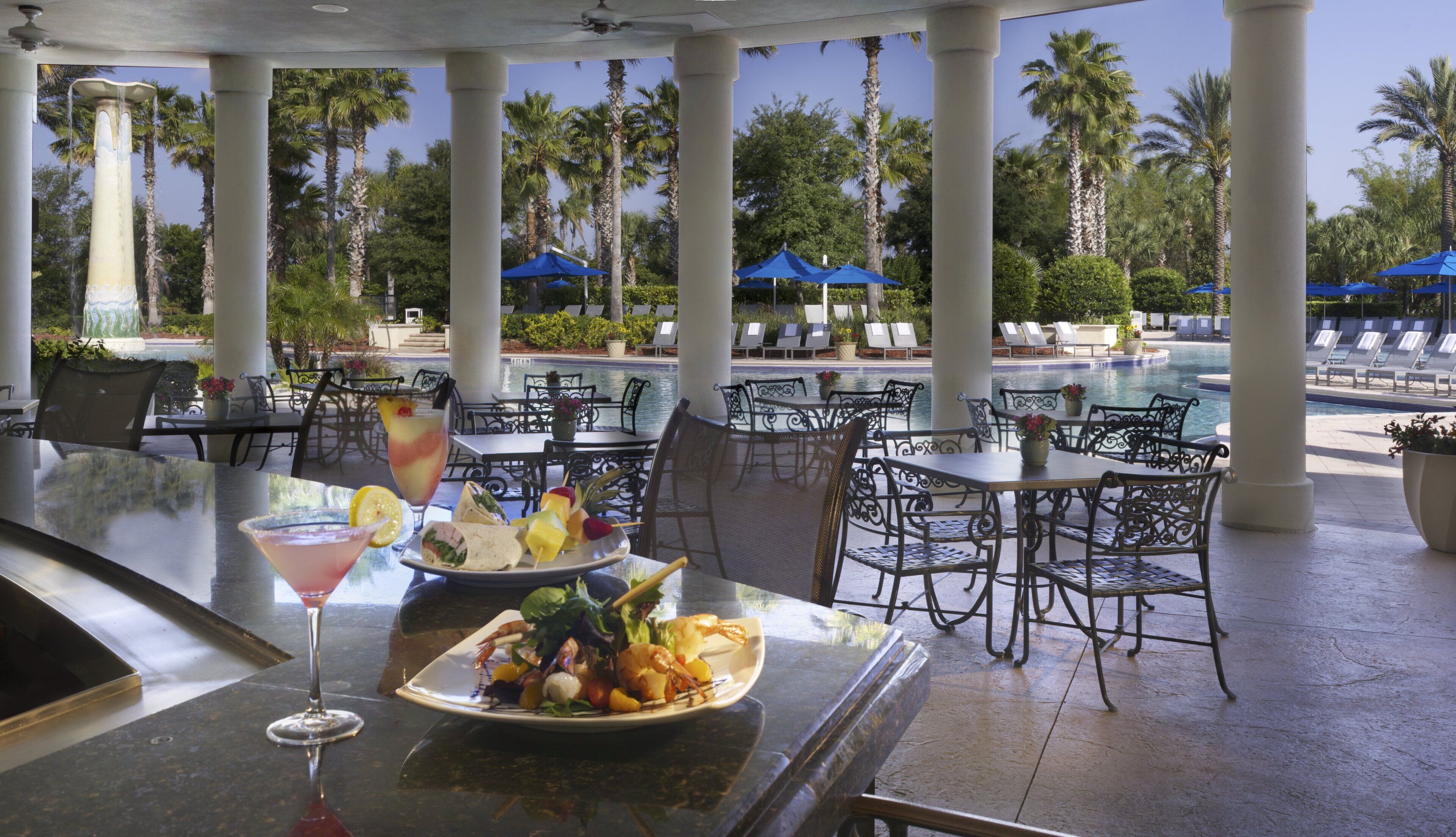 An outdoor dining area with tables and chairs, a pool, and palm trees in the background.