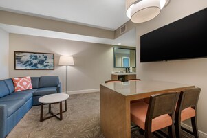 A hotel room with a sofa, a small table, a glass-top dining table with chairs, a flat-screen TV, a lamp, and a painting on the wall.