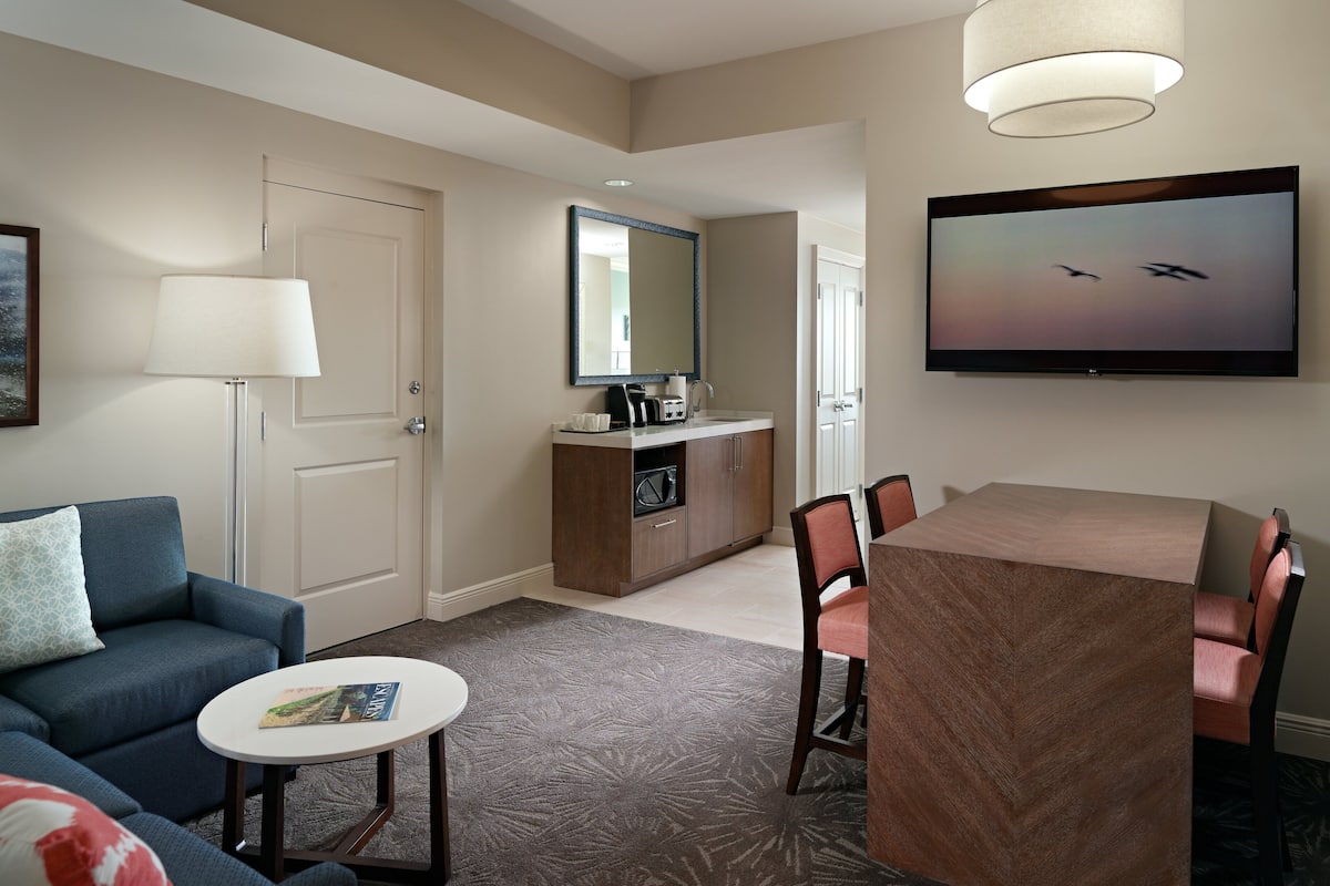 Ett hotellrum med en soffa, ett litet runt bord, en tv som är monterad på väggen, ett matbord med stolar och en minibar.