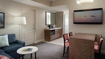 A hotel room with a sofa, a small round table, a TV mounted on the wall, a dining table with chairs, and a minibar.