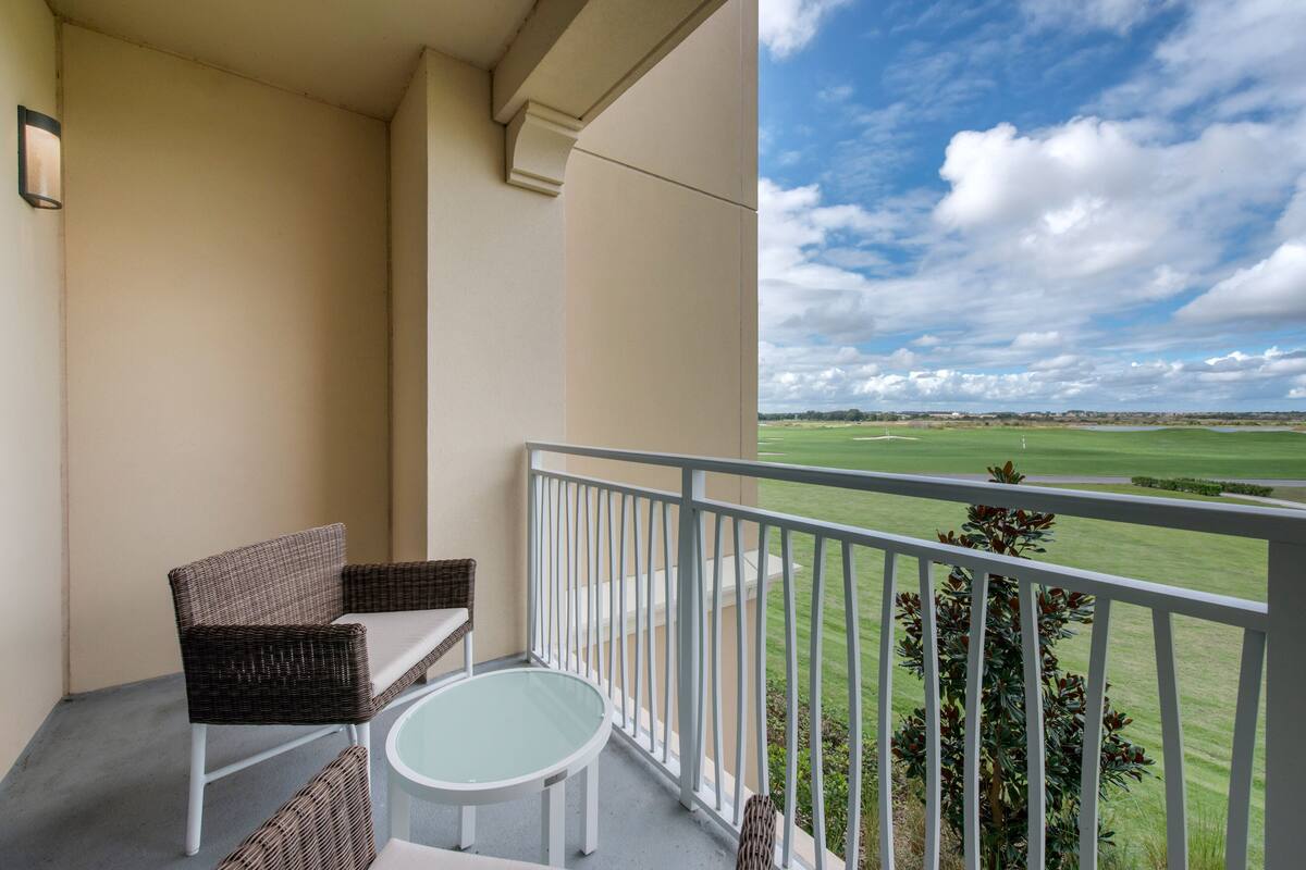 Un balcon doté de meubles en osier et d’une table en verre, offrant une vue sur un paysage verdoyant sous un ciel dégagé.