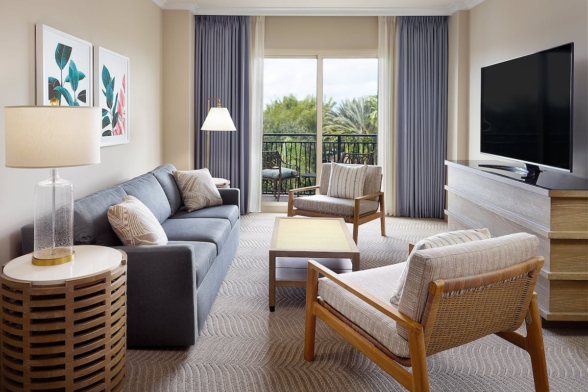 Une chambre d’hôtel moderne avec un canapé, deux fauteuils, une télévision et un balcon agrémenté de rideaux.