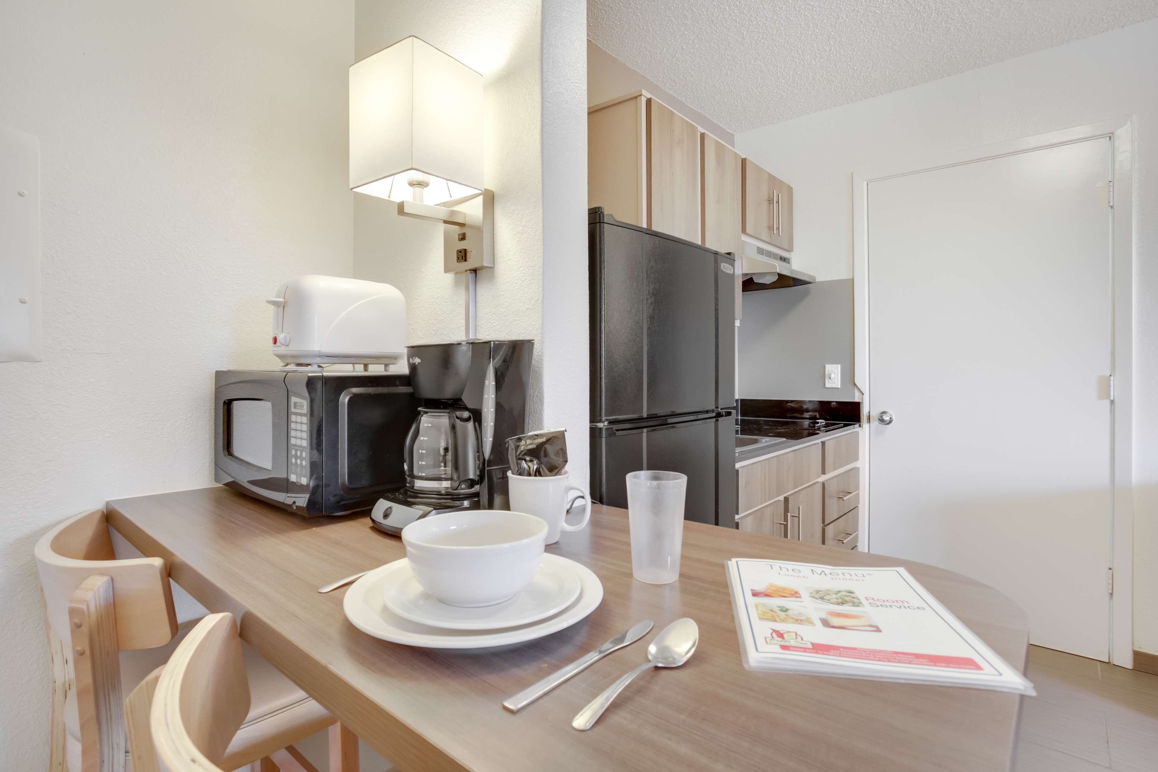 Una kitchenette compacta equipada con microondas, cafetera y refrigerador. Hay una mesa de comedor con un tazón blanco, una taza y cubiertos. Un menú está expuesto sobre la mesa.