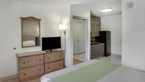 A hotel room with a bed, a TV on a wooden dresser, a mirror, a door leading to a kitchenette with a fridge and cabinets. - Studio 6 Stafford, TX - Houston - Sugarland (Stafford)