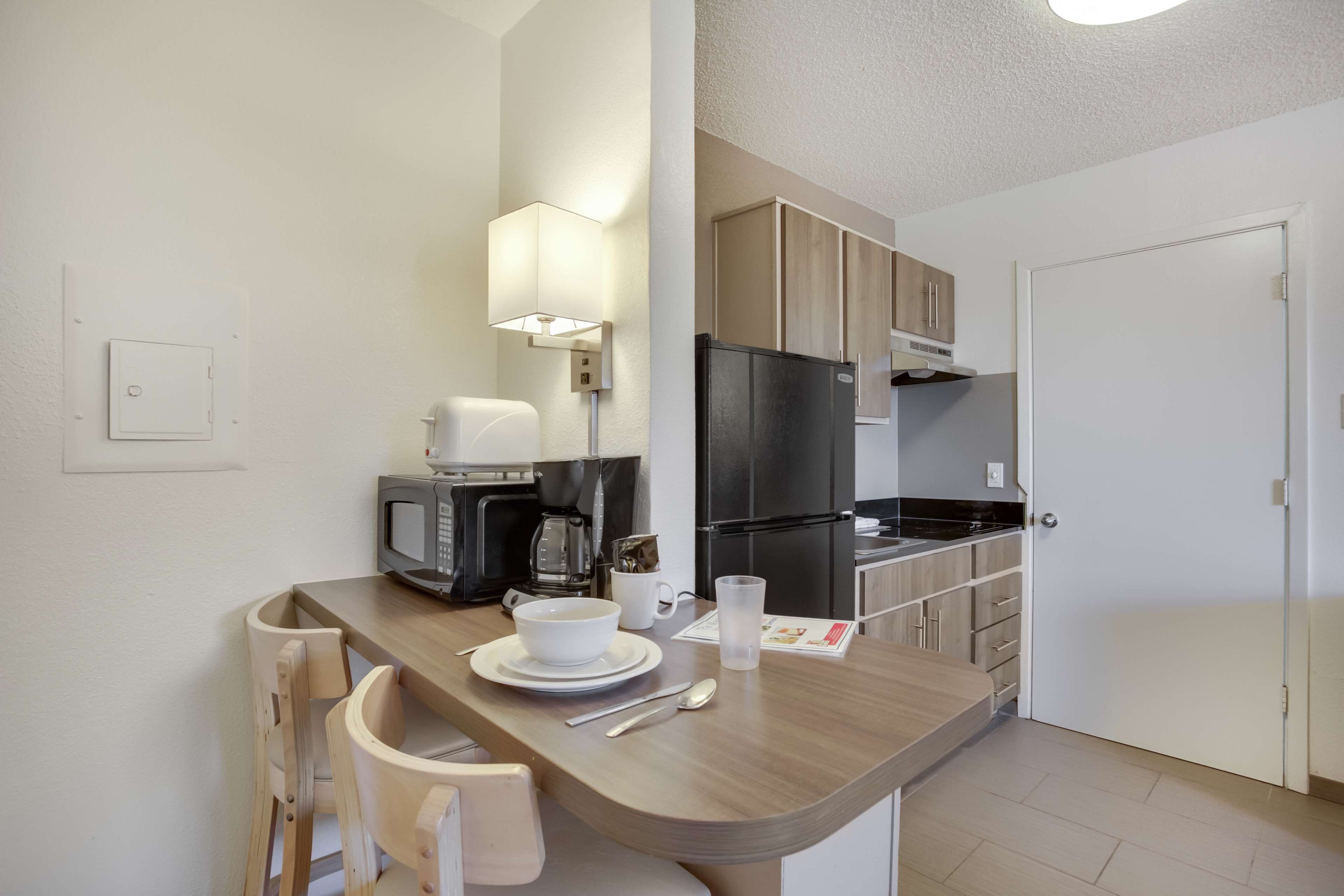 Cocina compacta equipada con microondas, cafetera y refrigerador. Cuenta con una mesa de comedor con sillas y un mueble pequeño.