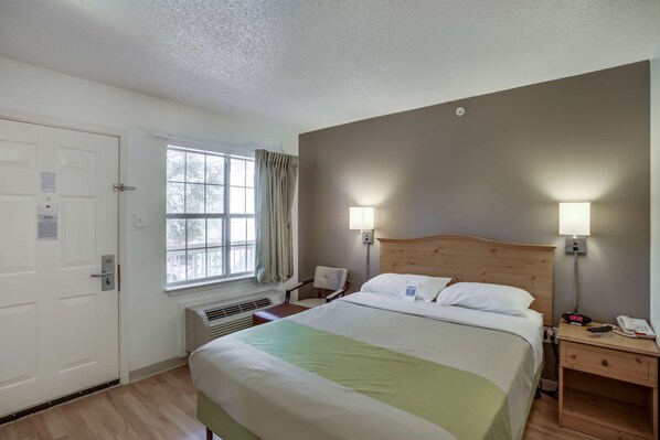 A hotel room with a bed, bedside lamps, a nightstand, a window with curtains, and a door. - Studio 6 Stafford, TX - Houston - Sugarland (Stafford)