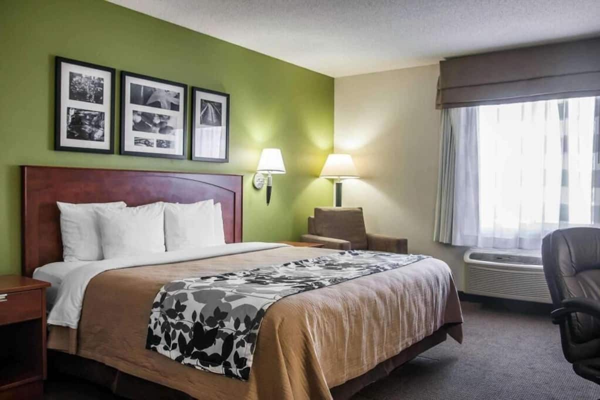 A hotel room with a bed, a chair, a desk, a lamp, and a window with curtains.