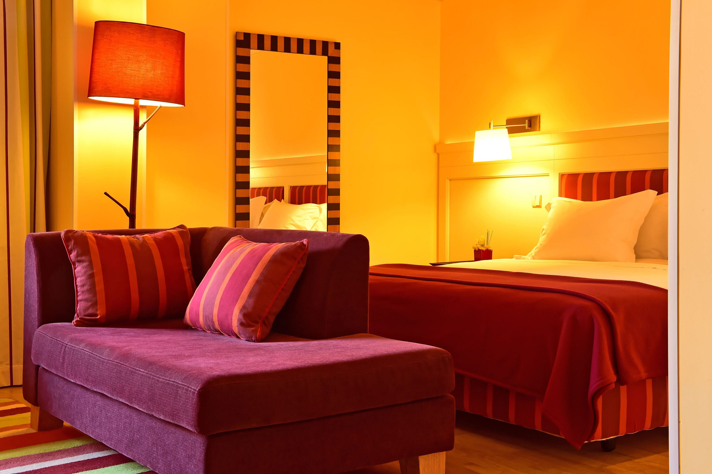 A hotel room with a purple sofa, a bed with red bedding, a large mirror, and a floor lamp.
