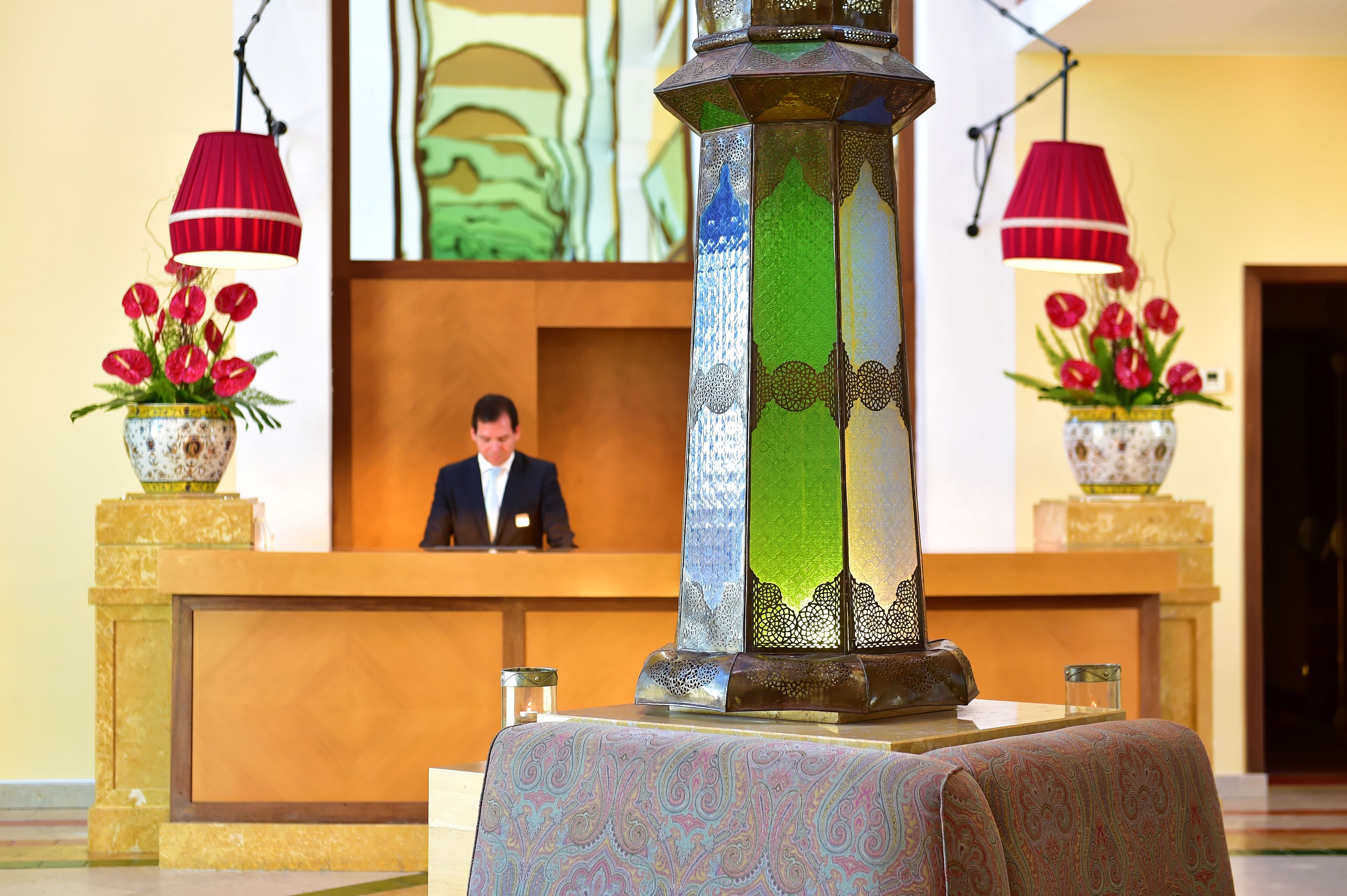 A reception desk with a man seated behind it, a decorative column, and two lamps with red shades.