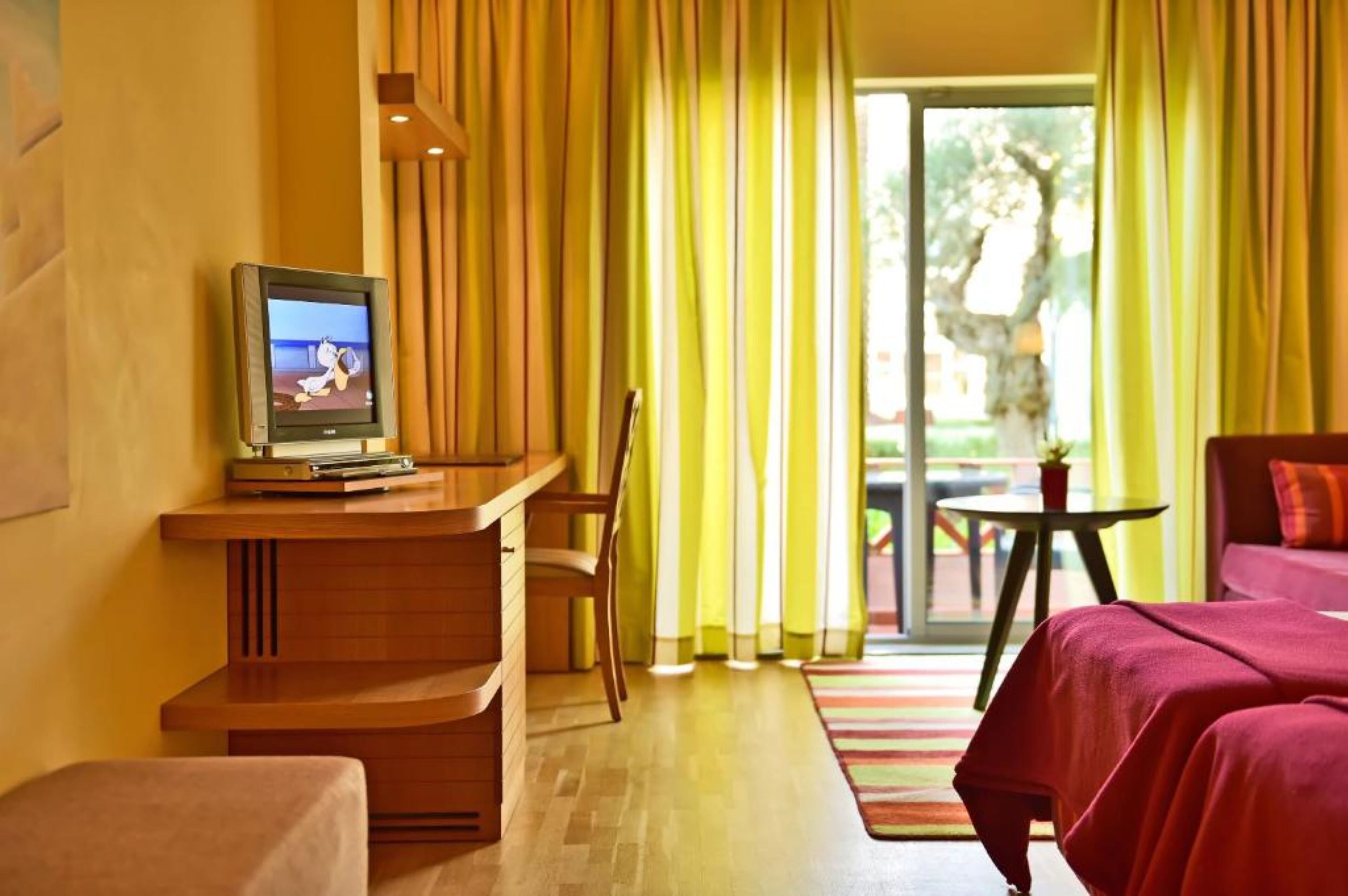 Une chambre d’hôtel avec une télévision posée sur un bureau en bois, une chaise, un lit avec une literie rouge et une vue sur la verdure à l’extérieur.