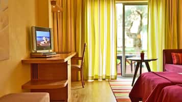 Une chambre d’hôtel avec une télévision posée sur un bureau en bois, une chaise, un lit avec une literie rouge et une vue sur la verdure à l’extérieur.
