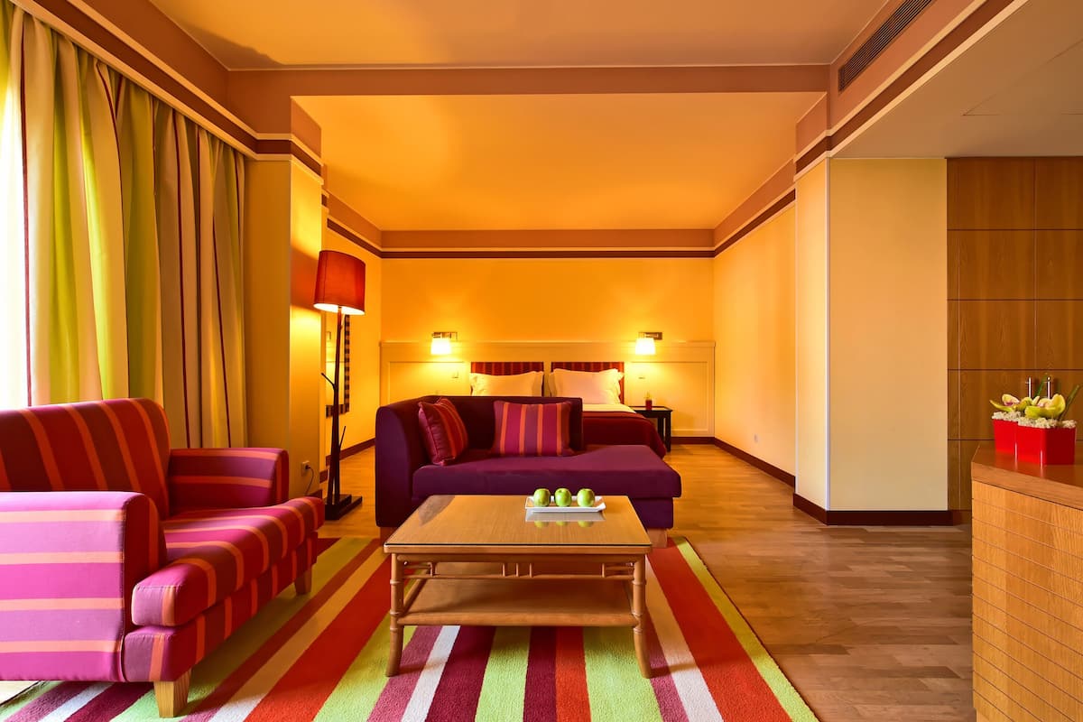 Quarto de hotel com sofá, poltrona e uma pequena mesa com maçãs. Há uma luminária e um armário de madeira com frutas.