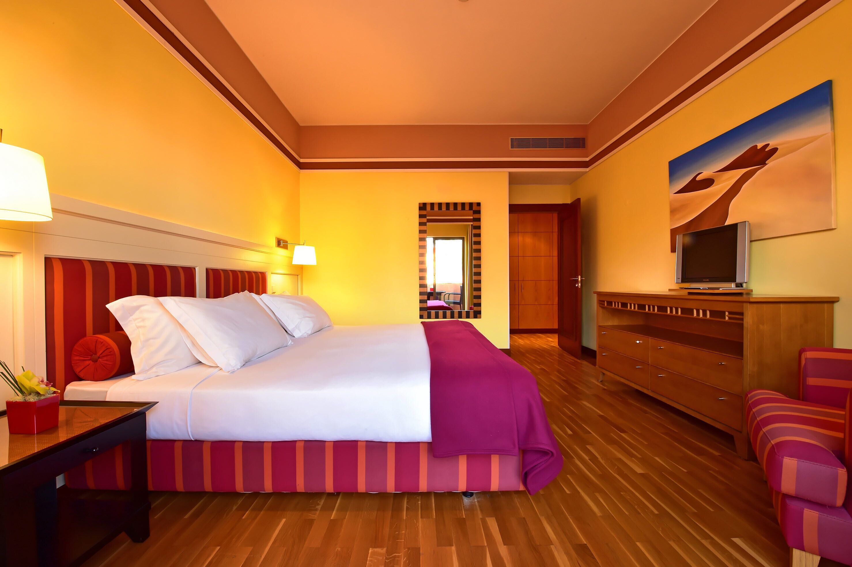 Habitación de hotel con cama, escritorio, televisor, cómoda y un cuadro en la pared.