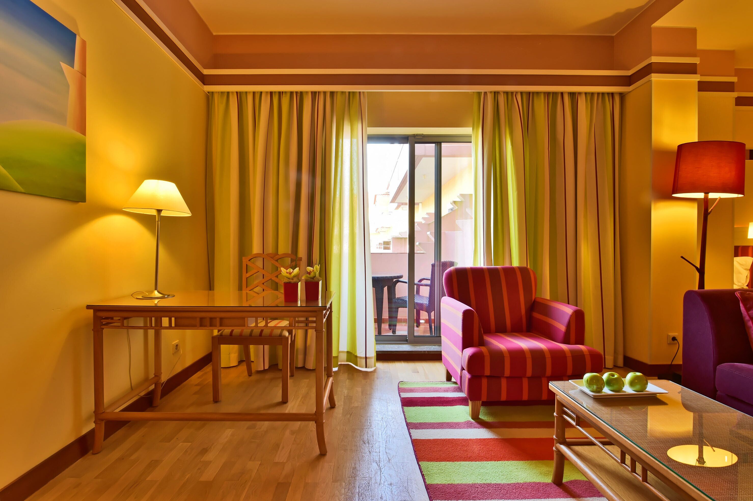 A hotel room with a wooden table, a striped armchair, a sofa, and a glass coffee table with apples.