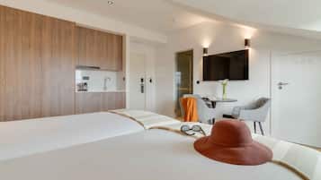 A modern hotel room with a large bed, a wooden wardrobe, a small table with a chair, a TV mounted on the wall, and a door leading to another room.