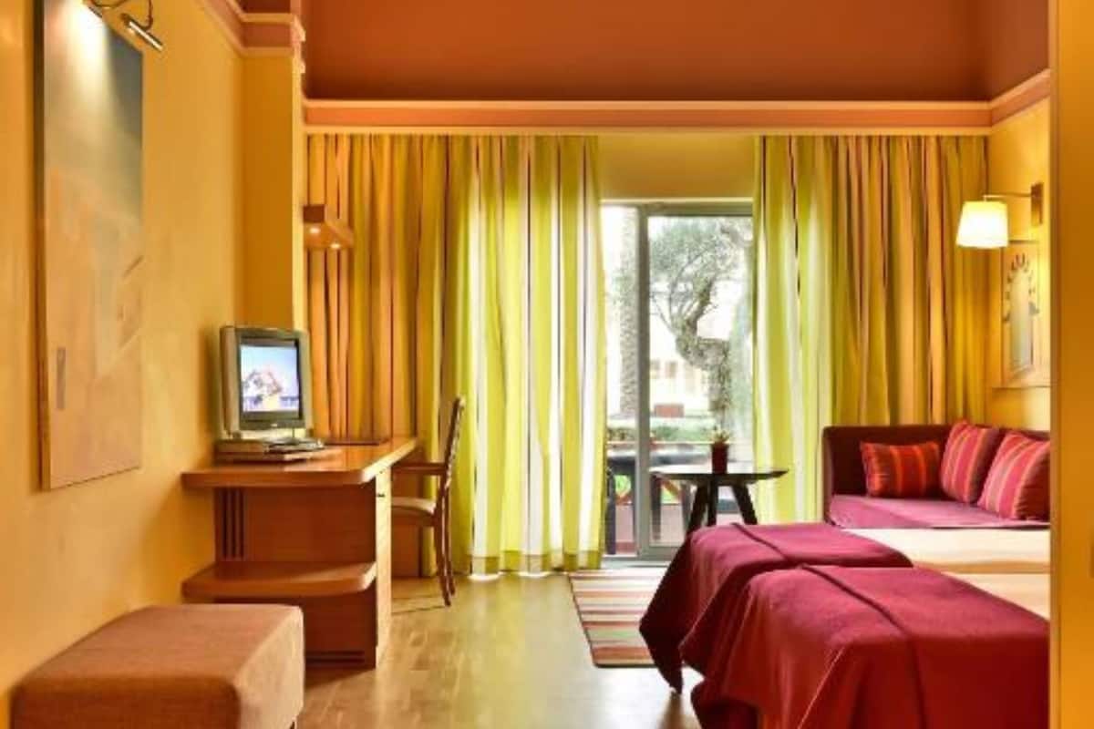 Quarto de hotel com piso de madeira, cama com roupa de cama vermelha, escrivaninha com televisão e janela com cortinas amarelas.