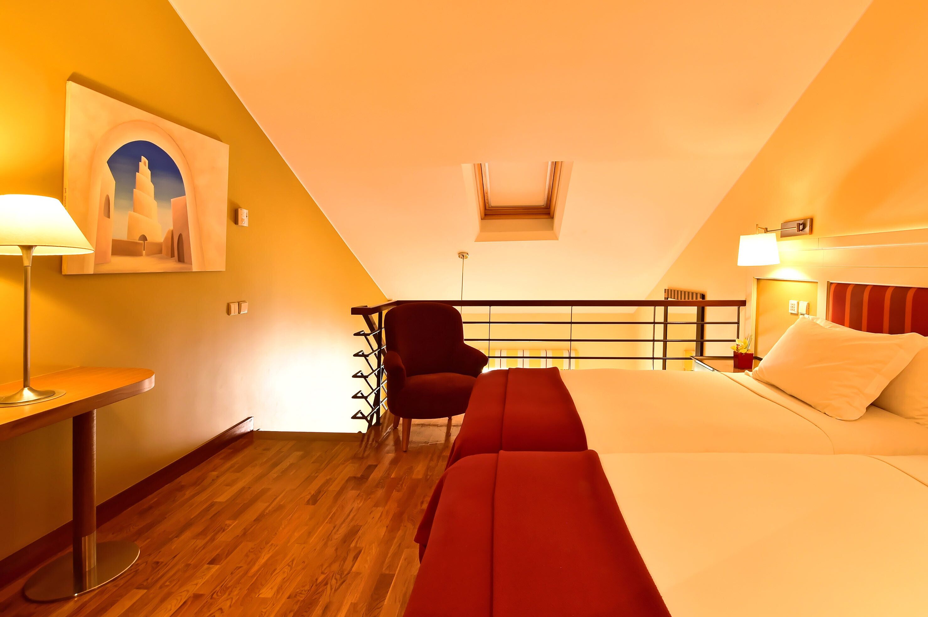 Een hotelkamer met een houten vloer, een bed met rode beddengoed, een bureau met een lamp, een stoel en een schilderij van een gebouw aan de muur.