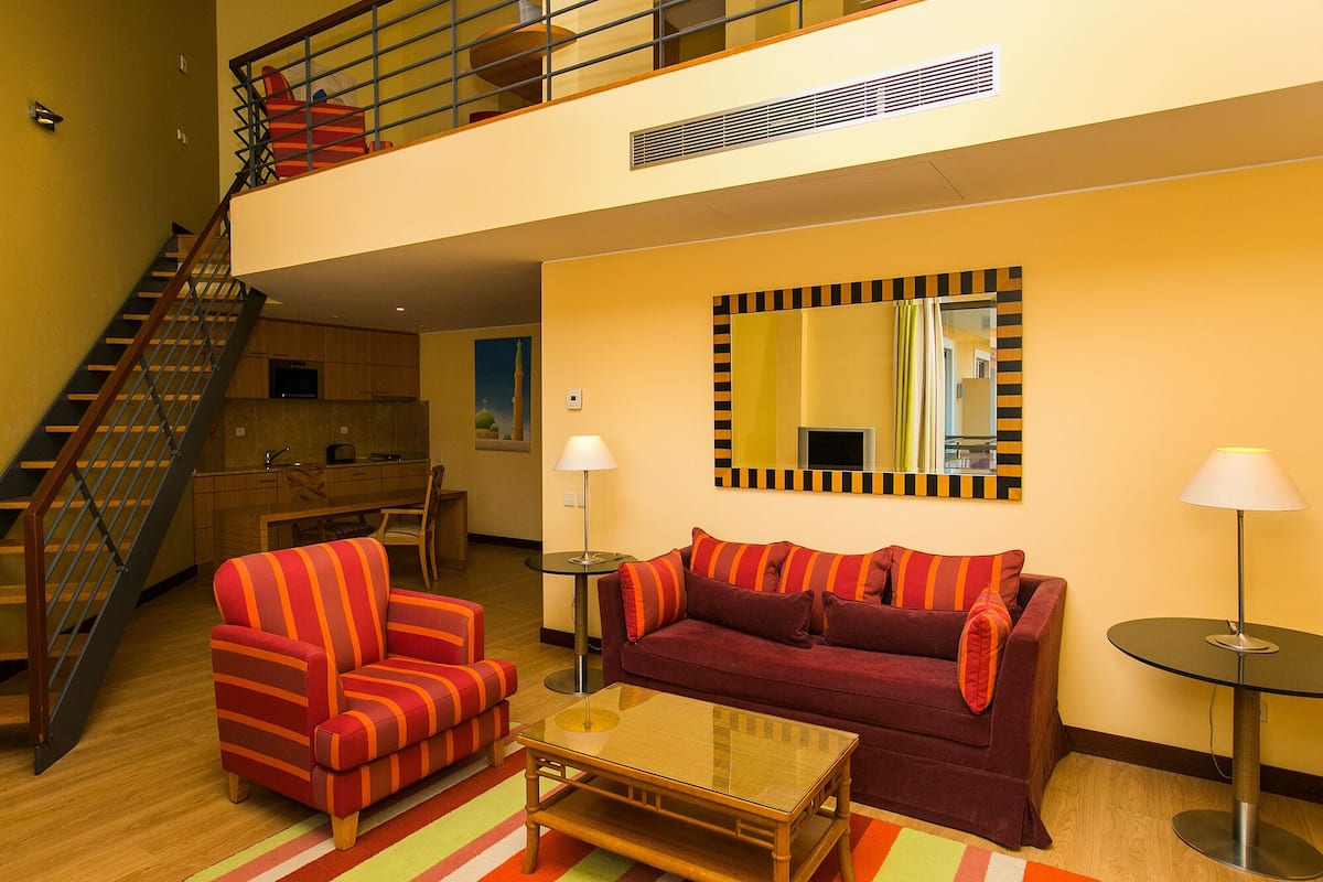 Uma sala de estar moderna com um sofá, poltrona e mesa de centro de vidro. Uma escada leva a um andar superior.