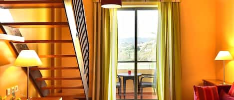 Una habitación de hotel moderna con una escalera de madera, un comedor con mesa y sillas, un amplio ventanal con cortinas y una alfombra colorida a rayas.