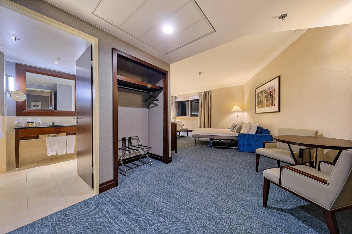 Habitación de hotel con baño, zona de estar con una mesa y sillas, y un espejo grande.