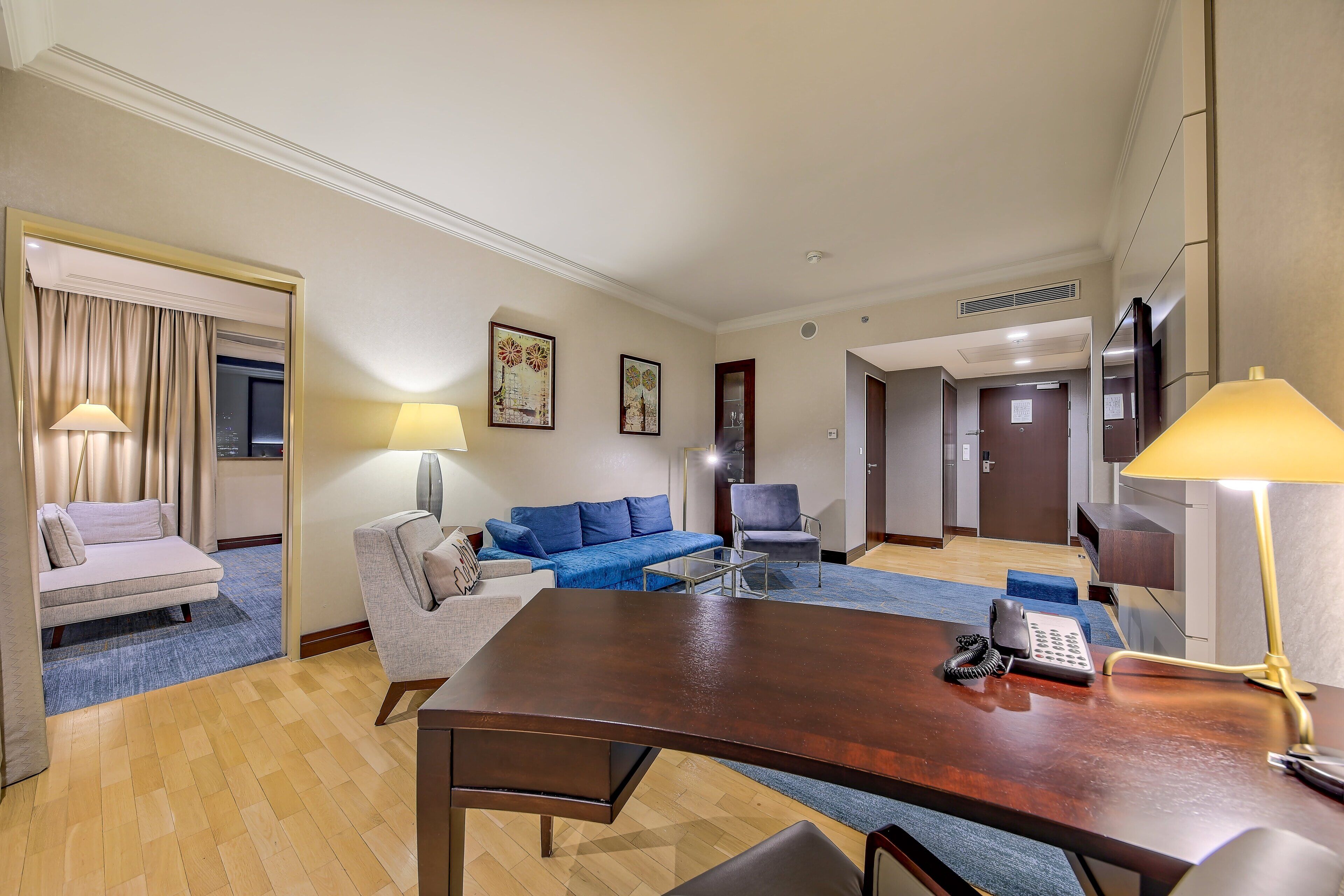 Een hotelkamer met een houten bureau, een blauwe bank, een grijze fauteuil, een klein bijzettafeltje en een lamp. Op het bureau staat een telefoon en er is een deur naar de gang.