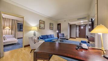 Een hotelkamer met een houten bureau, een blauwe bank, een grijze fauteuil, een klein bijzettafeltje en een lamp. Op het bureau staat een telefoon en er is een deur naar de gang.