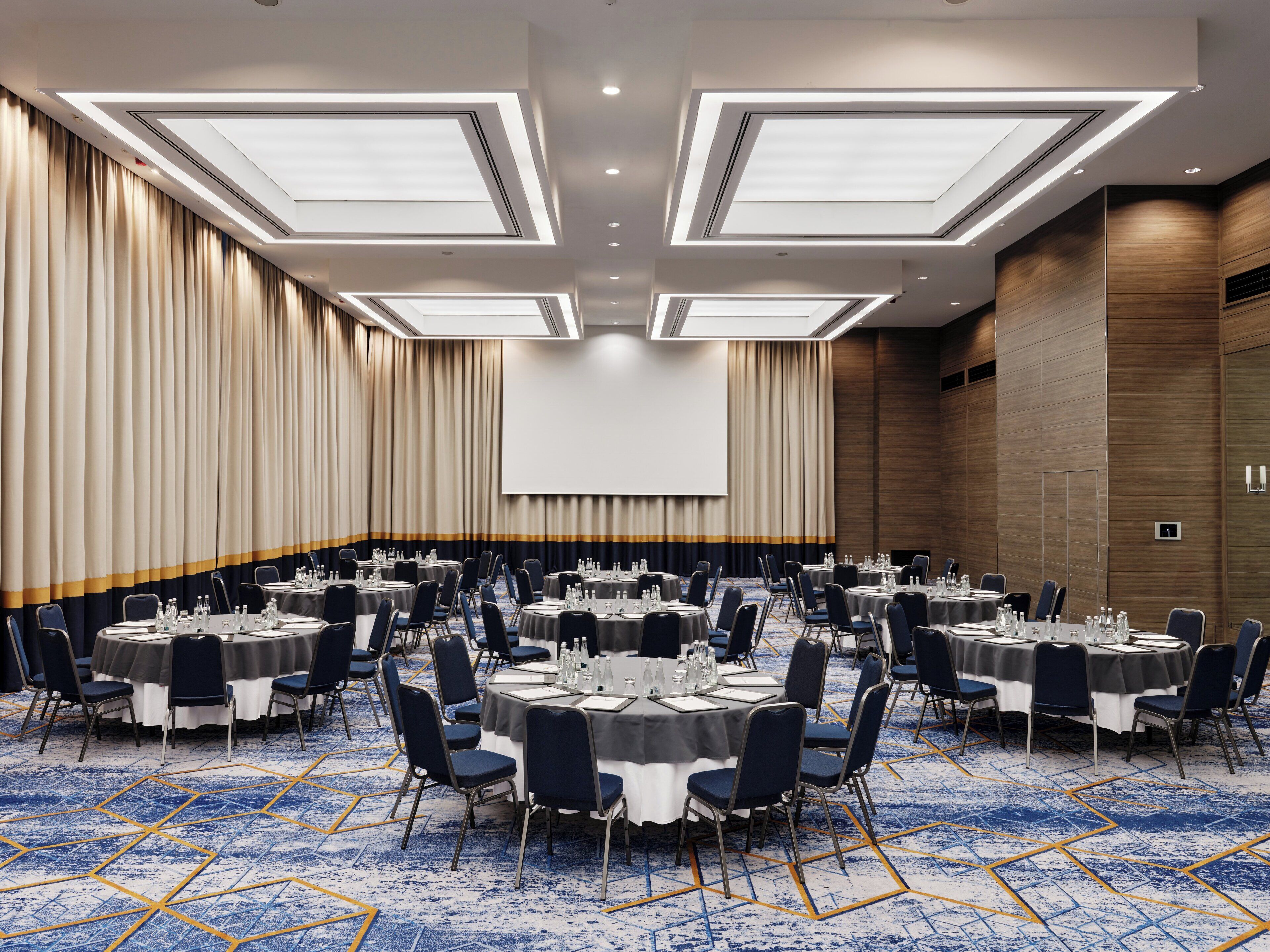 Un salón de conferencias con varias mesas y sillas, una pantalla grande y una alfombra estampada.