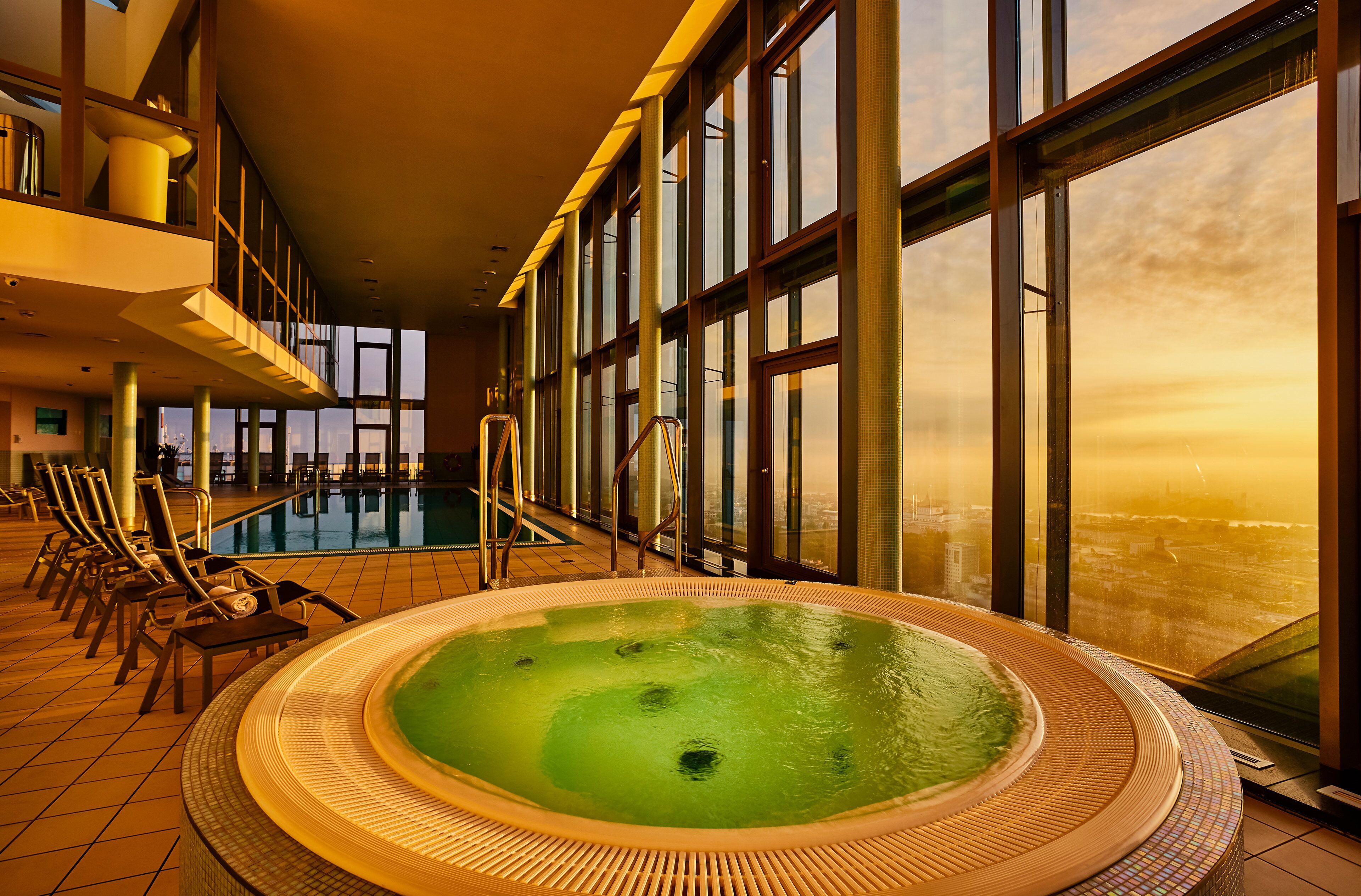 A large indoor swimming pool with a hot tub, surrounded by lounge chairs and large windows offering a city view.