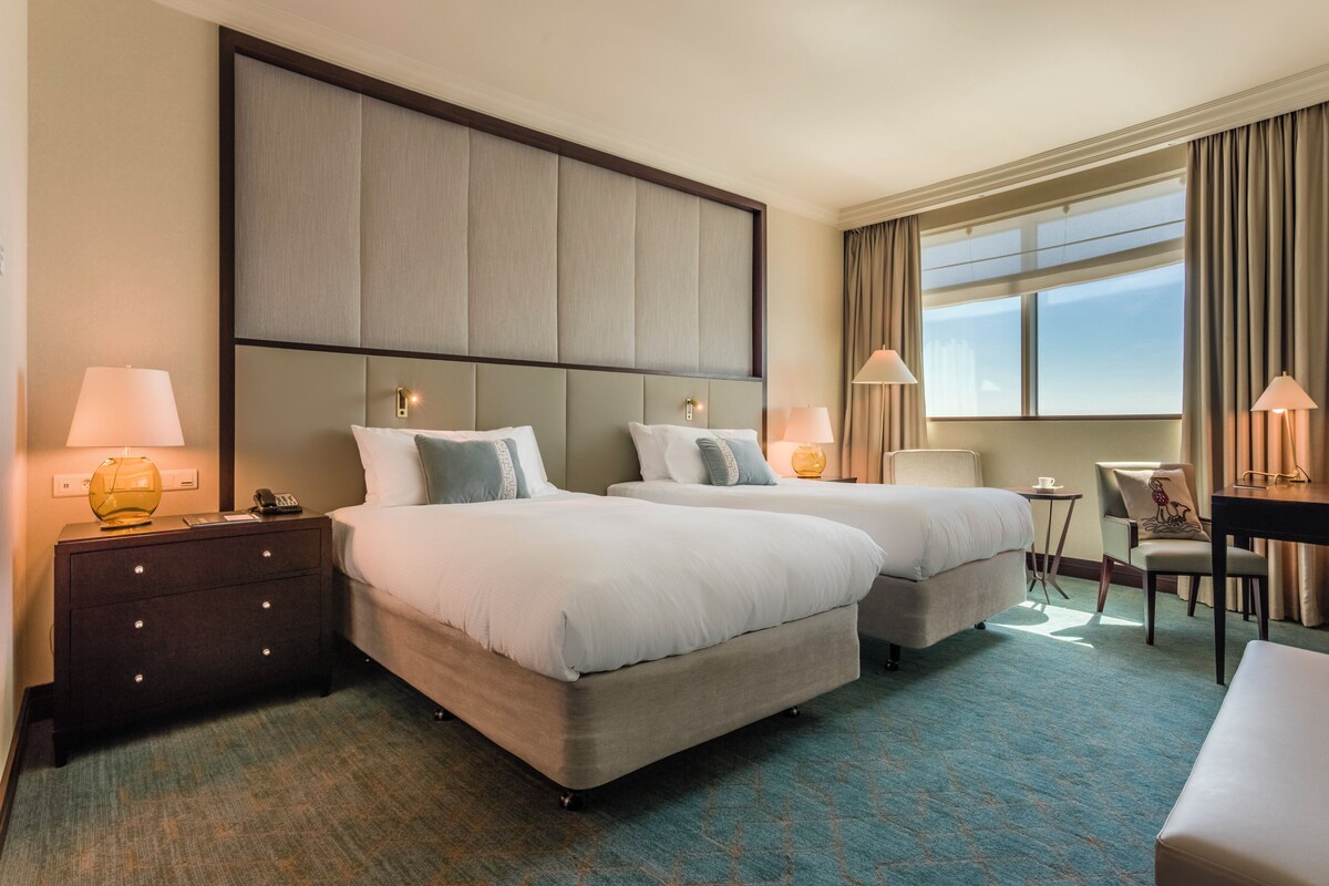 A hotel room with two beds, a desk, a chair, and a large window.