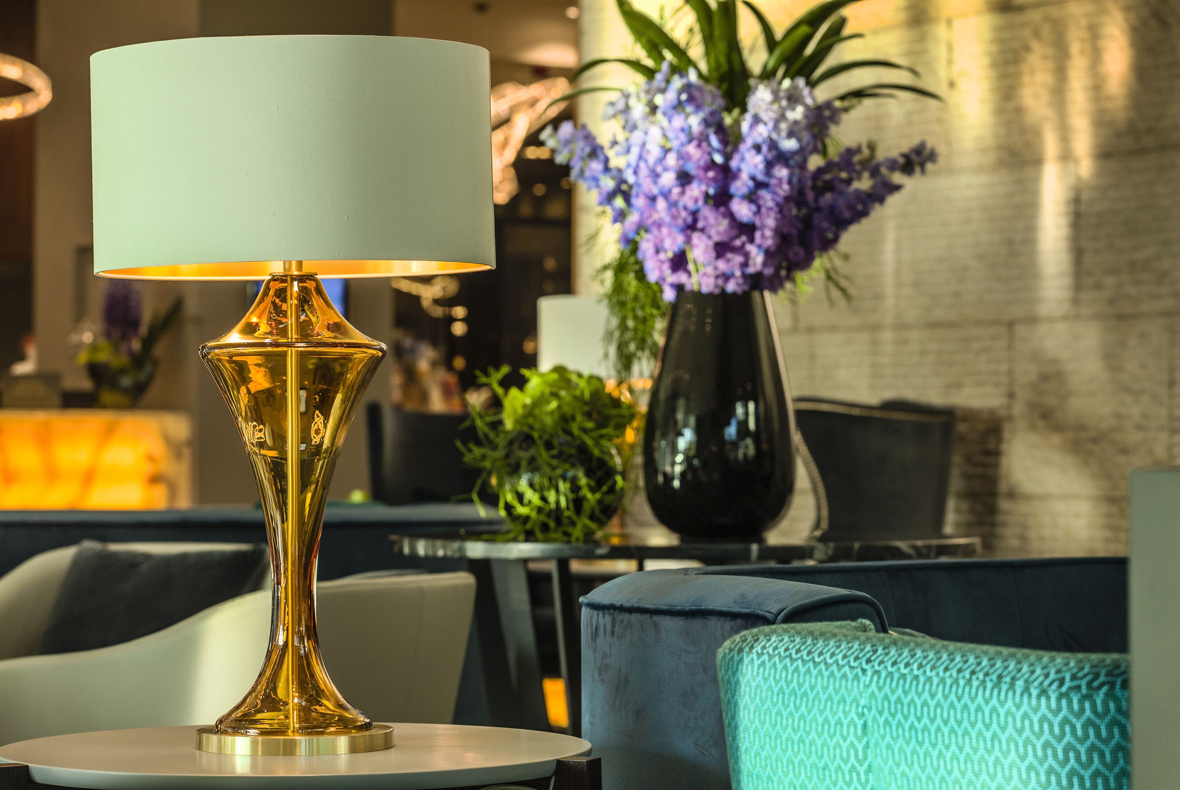 A modern lounge area with a gold lamp on a round table, a blue sofa, and a vase of purple flowers.