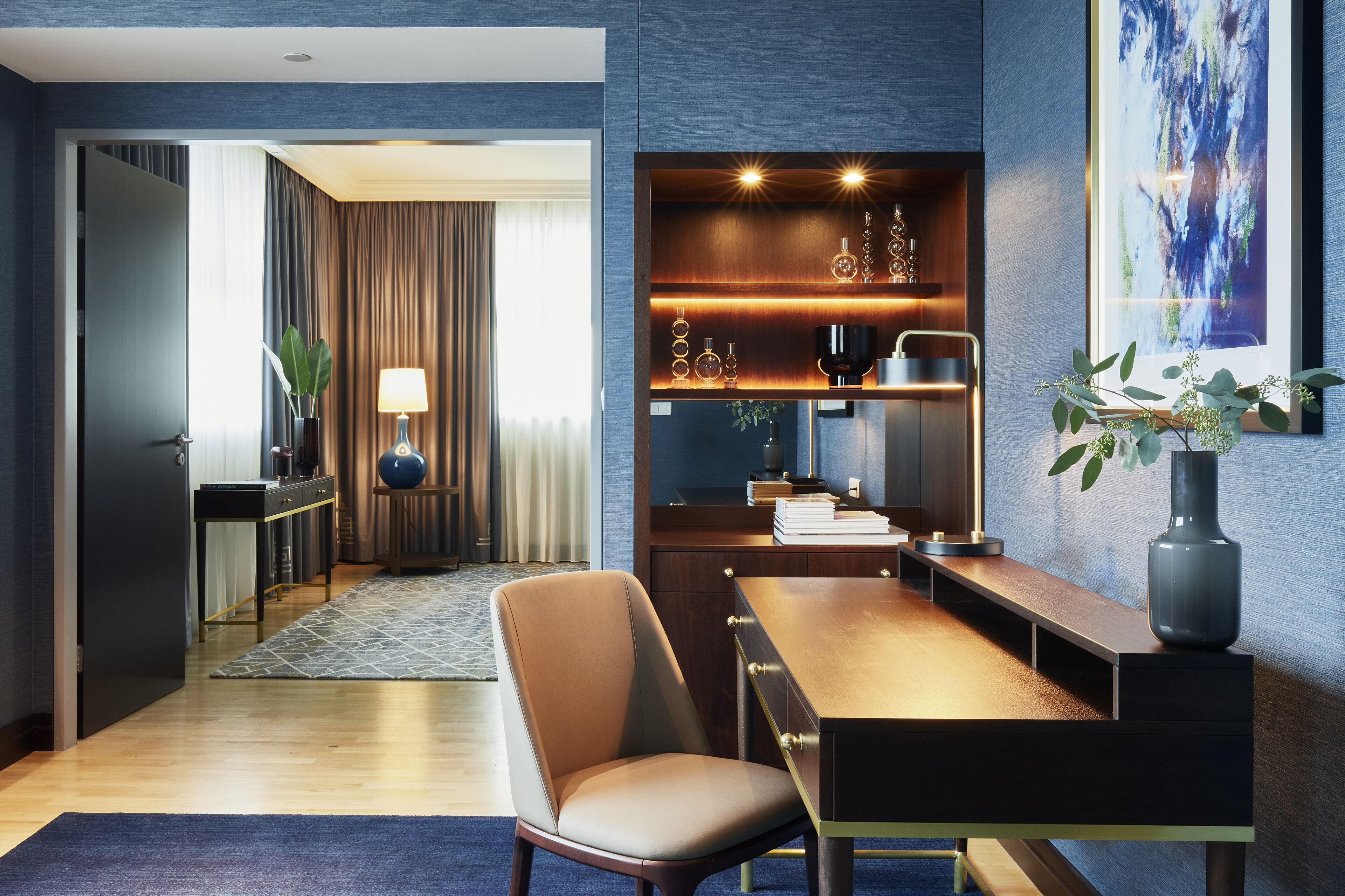 A modern hotel room with a wooden dining table, a comfortable chair, a small side table with a lamp, and a decorative shelf with glassware.