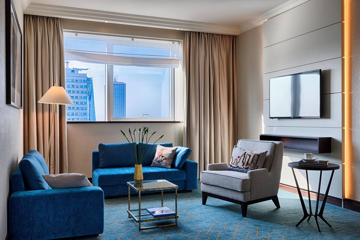 Una moderna camera d'hotel con un'ampia finestra, una televisione a schermo piatto, un divano blu, una poltrona grigia e un piccolo tavolino.