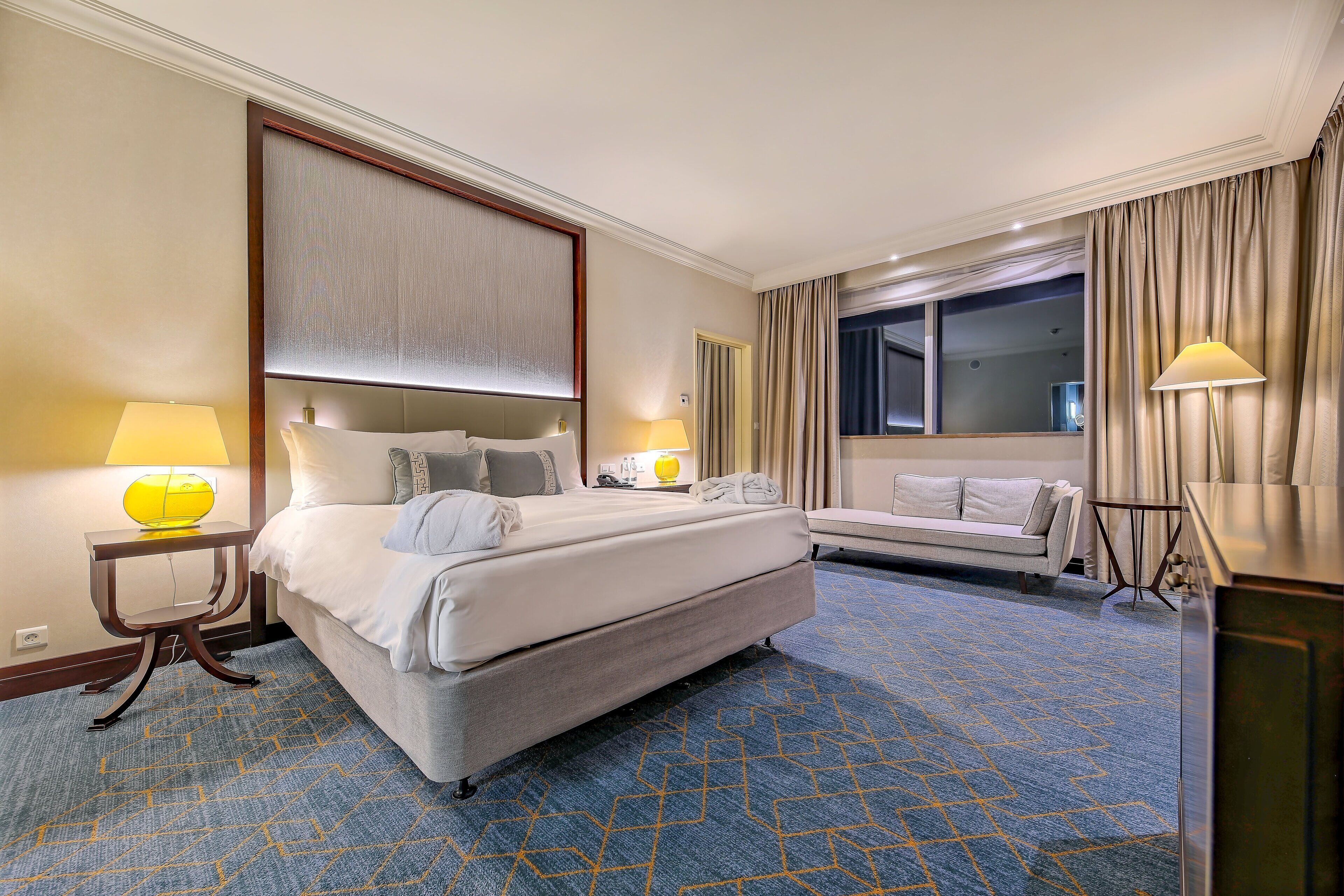 A hotel room with a large bed, bedside tables with lamps, a sofa, a desk, and a large window with curtains.