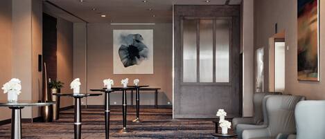 Un vestíbulo de hotel con una alfombra azul estampada, una zona de estar con una silla gris, una mesa con un jarrón de flores y un cuadro abstracto de gran tamaño en la pared.