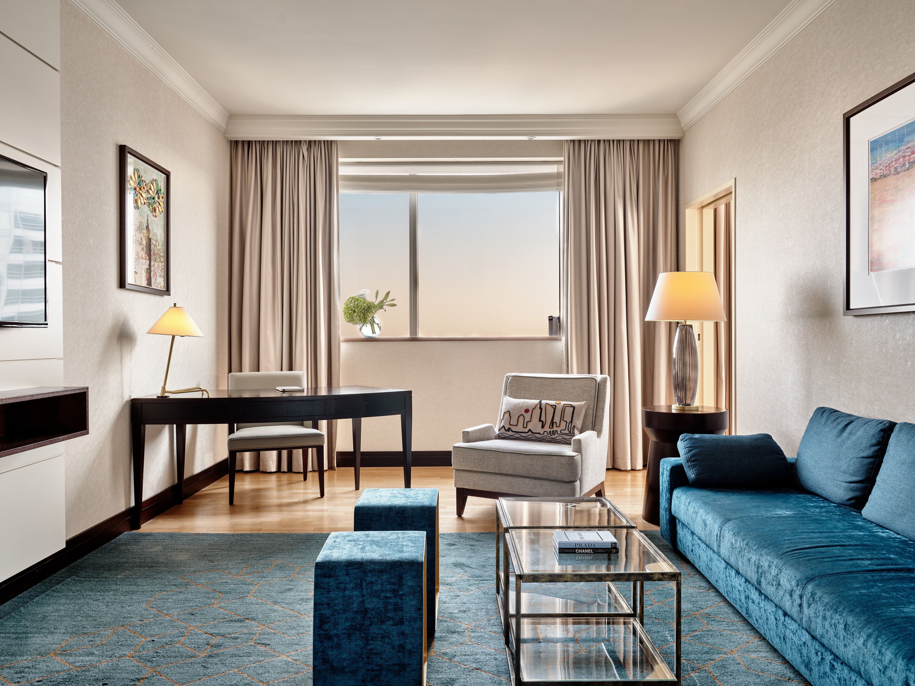 A modern hotel room with a sofa, armchair, coffee table, and a desk with a lamp.