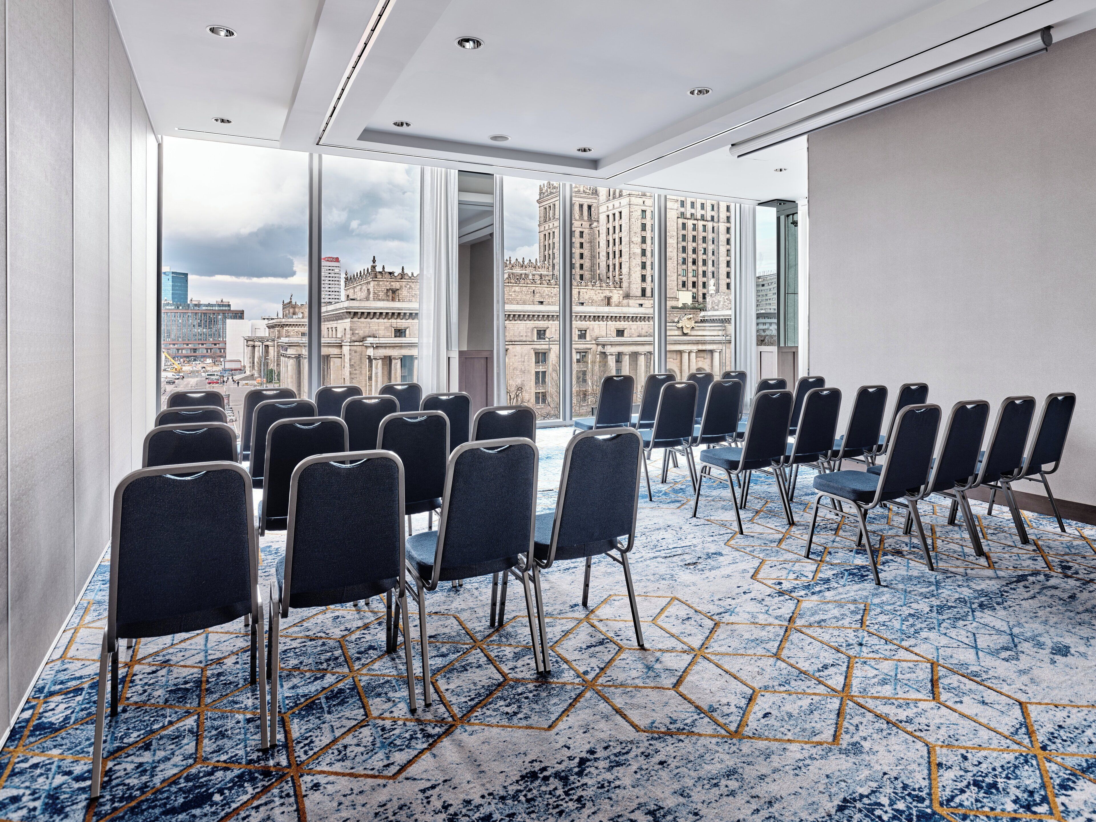 A conference room with rows of chairs, large windows with a city view, and a patterned carpet.