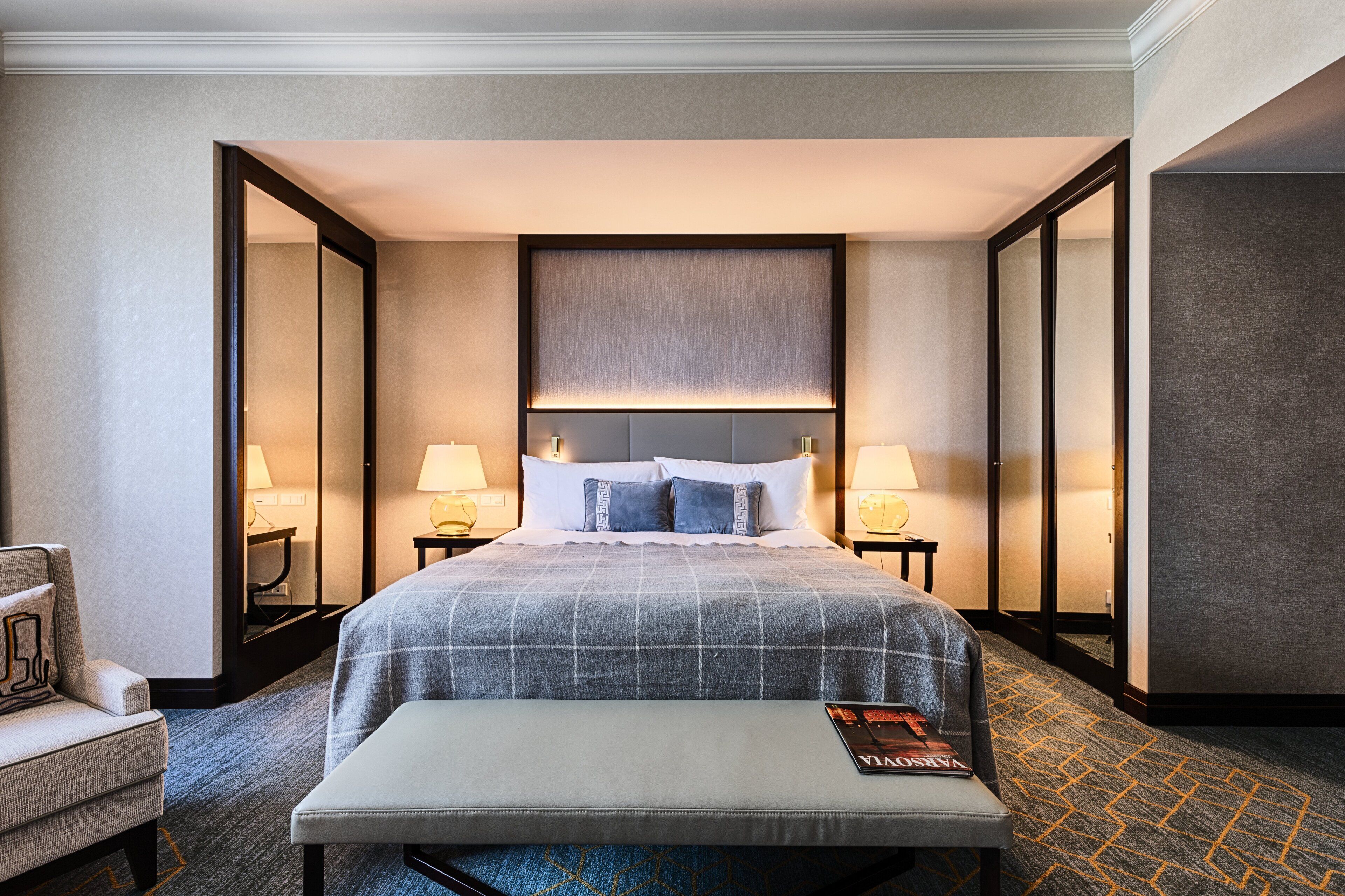 A hotel room with a large bed, two bedside tables with lamps, a bench, and a mirror.