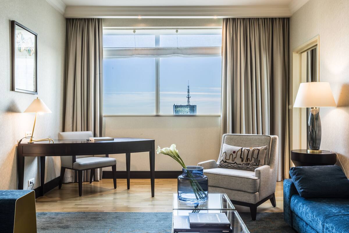 Una habitación de hotel moderna con una mesa de comedor, un sofá, una silla, una mesita de centro con un jarrón y vistas a un paisaje urbano a través de la ventana.
