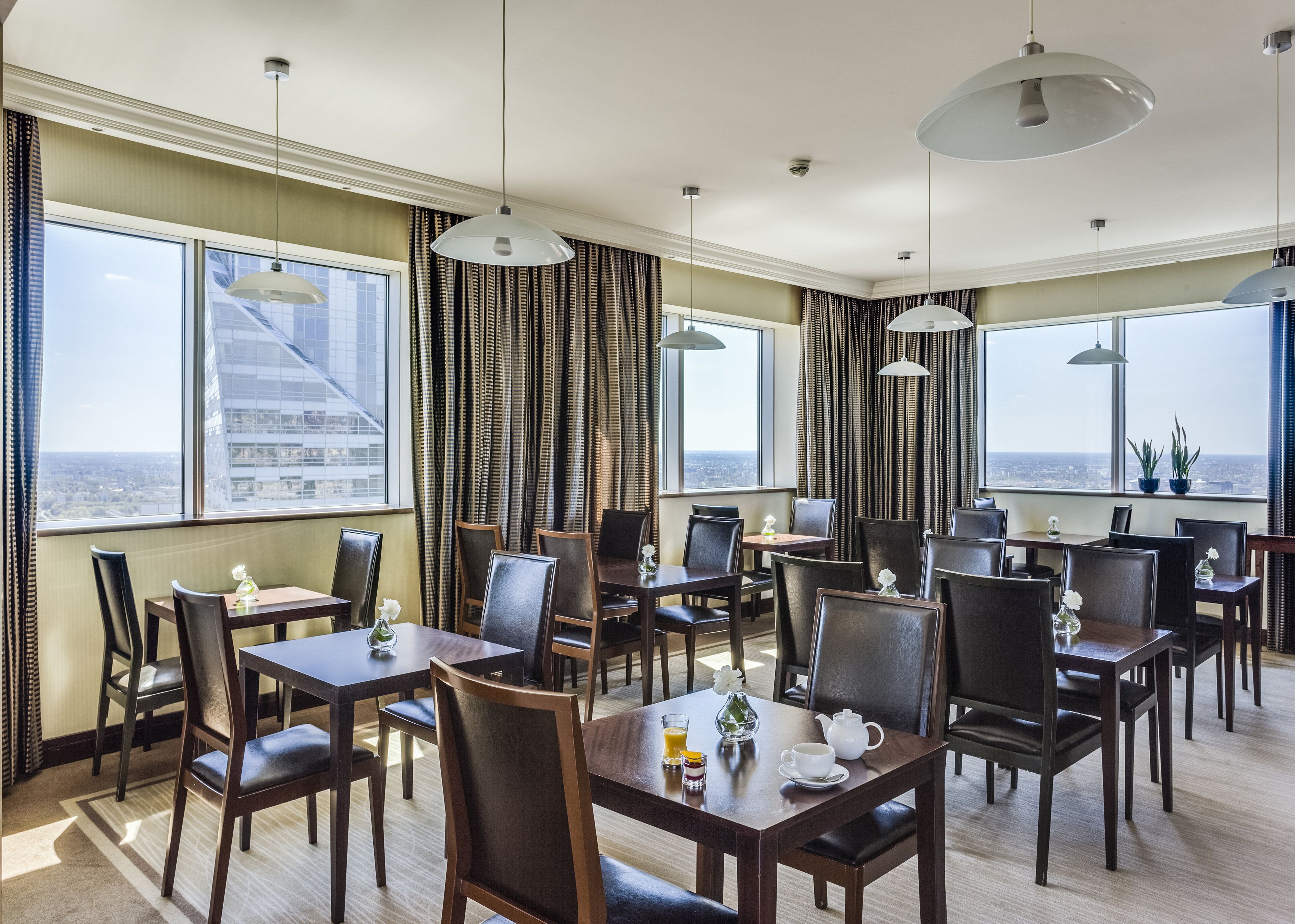 A hotel dining area with multiple tables and chairs, large windows with city views, and modern pendant lighting.
